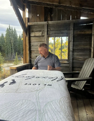 A man dressed in a gray shirt sits on a bed, surrounded by a calm and inviting atmosphere.